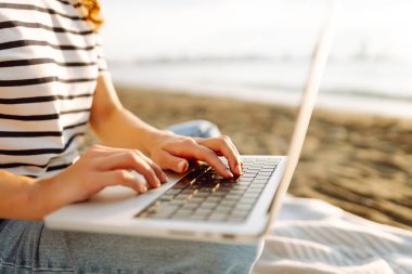 Güneşli bir günde plaj battaniyesinde otururken dizüstü bilgisayarla çalışan serbest çalışan bir kadın. Dizüstü bilgisayarı olan güzel bir kadın gün batımında doğanın ve deniz manzarasının tadını çıkarıyor. Rahatlama kavramı, dinlenme.