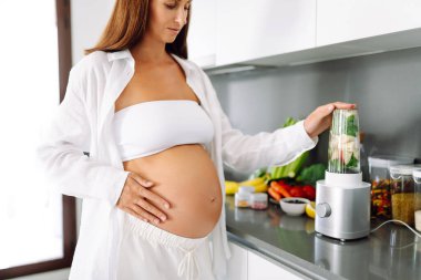 Hamile kadın modern bir mutfakta yeşil meyve püresi hazırlıyor. Bebek bekleyen genç bir kadın sağlığına dikkat eder ve sağlıklı bir içecek hazırlamak için blender kullanır. Sağlık ve gıda kavramı.