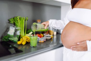 Hamile kadın modern bir mutfakta yeşil meyve püresi hazırlıyor. Bebek bekleyen genç bir kadın sağlığına dikkat eder ve sağlıklı bir içecek hazırlamak için blender kullanır. Sağlık ve gıda kavramı.