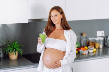 Hamile kadın modern bir mutfakta yeşil meyve püresi hazırlıyor. Bebek bekleyen genç bir kadın sağlığına dikkat eder ve sağlıklı bir içecek hazırlamak için blender kullanır. Sağlık ve gıda kavramı.