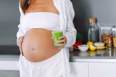 Hamile kadın modern bir mutfakta yeşil meyve püresi hazırlıyor. Bebek bekleyen genç bir kadın sağlığına dikkat eder ve sağlıklı bir içecek hazırlamak için blender kullanır. Sağlık ve gıda kavramı.