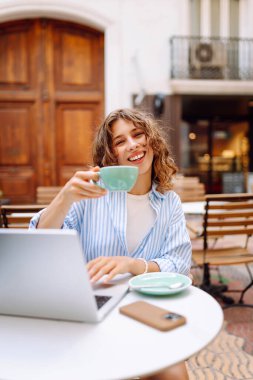 Dizüstü bilgisayarı olan serbest çalışan genç bir bayan sıcak içecekle birlikte terasta çalışıyor. Laptoplu neşeli kadın güneşli günün ve açık havada kahvenin tadını çıkarıyor. Serbest çalışma, teknoloji ve boş zaman kavramları.