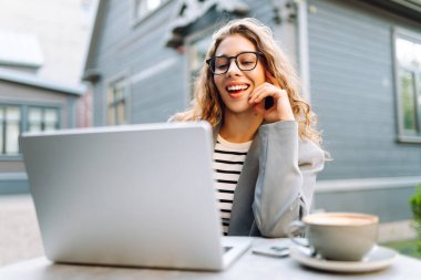 Dizüstü bilgisayarı olan neşeli bir kadın kafe terasında bir masada oturuyor. Dişi bir serbest çalışan dizüstü bilgisayarında çalışır, kahve içer ve güneşli günün tadını çıkarır. Uzaktan çalışma konsepti, hafta sonu.