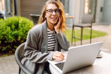 Dizüstü bilgisayarı olan neşeli bir kadın kafe terasında bir masada oturuyor. Dişi bir serbest çalışan dizüstü bilgisayarında çalışır, kahve içer ve güneşli günün tadını çıkarır. Uzaktan çalışma konsepti, hafta sonu.