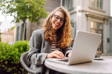 Dizüstü bilgisayarı olan neşeli bir kadın kafe terasında bir masada oturuyor. Dişi bir serbest çalışan dizüstü bilgisayarında çalışır, kahve içer ve güneşli günün tadını çıkarır. Uzaktan çalışma konsepti, hafta sonu.