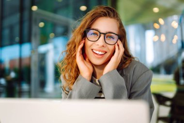 Dizüstü bilgisayarı olan neşeli bir kadın kafe terasında bir masada oturuyor. Dişi bir serbest çalışan dizüstü bilgisayarında çalışır, kahve içer ve güneşli günün tadını çıkarır. Uzaktan çalışma konsepti, hafta sonu.