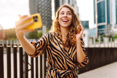 Mutlu bir kadın telefonuyla modern bir şehrin arka planına selfie çeker. Telefon blogları olan güzel bir turist ve gün batımının tadını çıkarıyor. Blogculuk, boş zaman ve kentsellik kavramları.