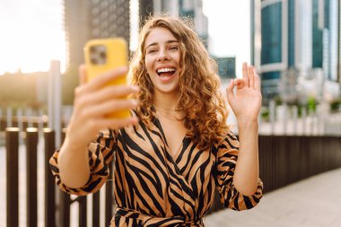 Mutlu bir kadın telefonuyla modern bir şehrin arka planına selfie çeker. Telefon blogları olan güzel bir turist ve gün batımının tadını çıkarıyor. Blogculuk, boş zaman ve kentsellik kavramları.