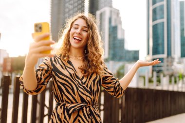 Mutlu bir kadın telefonuyla modern bir şehrin arka planına selfie çeker. Telefon blogları olan güzel bir turist ve gün batımının tadını çıkarıyor. Blogculuk, boş zaman ve kentsellik kavramları.