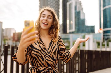 Mutlu bir kadın telefonuyla modern bir şehrin arka planına selfie çeker. Telefon blogları olan güzel bir turist ve gün batımının tadını çıkarıyor. Blogculuk, boş zaman ve kentsellik kavramları.
