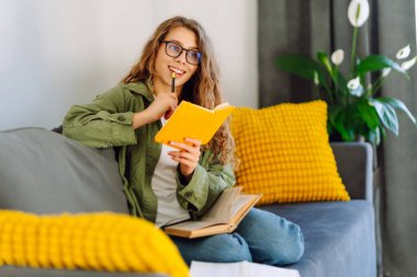 Sarı defterde gözlüklü güzel bir kadının portresi rahat bir odada kanepede otururken. Genç öğrenci rahatlar, evde notlar alır. Eğitim, rahatlama ve konfor kavramı.