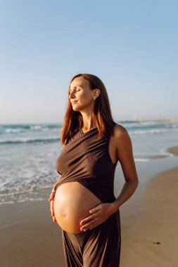 Çocuk bekleyen güzel bir kadın kumlu bir sahilde duruyor ve karnına dokunuyor. Hamile kadın güneşli günün ve deniz manzarasının tadını çıkarıyor. Huzur, hamilelik ve doğa kavramı.