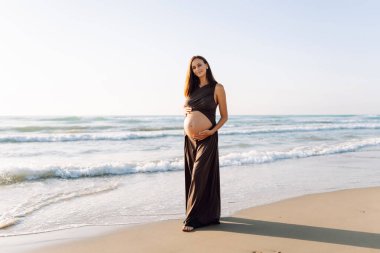 Çocuk bekleyen güzel bir kadın kumlu bir sahilde duruyor ve karnına dokunuyor. Hamile kadın güneşli günün ve deniz manzarasının tadını çıkarıyor. Huzur, hamilelik ve doğa kavramı.