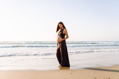 Çocuk bekleyen güzel bir kadın kumlu bir sahilde duruyor ve karnına dokunuyor. Hamile kadın güneşli günün ve deniz manzarasının tadını çıkarıyor. Huzur, hamilelik ve doğa kavramı.