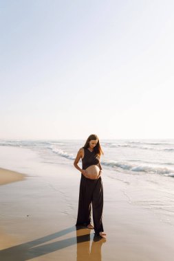 Çocuk bekleyen güzel bir kadın kumlu bir sahilde duruyor ve karnına dokunuyor. Hamile kadın güneşli günün ve deniz manzarasının tadını çıkarıyor. Huzur, hamilelik ve doğa kavramı.