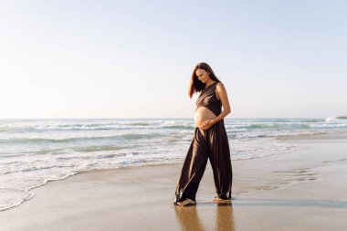 Çocuk bekleyen güzel bir kadın kumlu bir sahilde duruyor ve karnına dokunuyor. Hamile kadın güneşli günün ve deniz manzarasının tadını çıkarıyor. Huzur, hamilelik ve doğa kavramı.