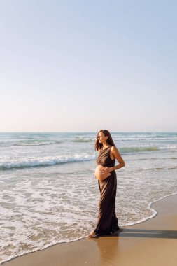 Çocuk bekleyen güzel bir kadın kumlu bir sahilde duruyor ve karnına dokunuyor. Hamile kadın güneşli günün ve deniz manzarasının tadını çıkarıyor. Huzur, hamilelik ve doğa kavramı.