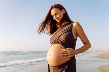 Çocuk bekleyen güzel bir kadın kumlu bir sahilde duruyor ve karnına dokunuyor. Hamile kadın güneşli günün ve deniz manzarasının tadını çıkarıyor. Huzur, hamilelik ve doğa kavramı.