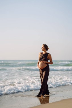 Çocuk bekleyen güzel bir kadın kumlu bir sahilde duruyor ve karnına dokunuyor. Hamile kadın güneşli günün ve deniz manzarasının tadını çıkarıyor. Huzur, hamilelik ve doğa kavramı.