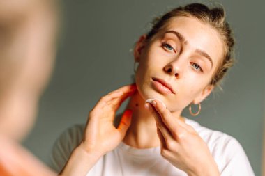 Genç bir kadının aynaya bakarken yüzüne pamuk pedlerle makyaj yaptığı yakın plan. Sıcak bir odada yüzünü önemseyen güzel bir kadın. Güzellik ve cilt bakımı, cilt bakımı.