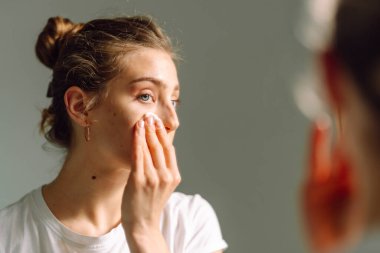Genç bir kadının aynaya bakarken yüzüne pamuk pedlerle makyaj yaptığı yakın plan. Sıcak bir odada yüzünü önemseyen güzel bir kadın. Güzellik ve cilt bakımı, cilt bakımı.