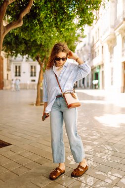 Şık giyinmiş genç bir kadın şehrin güneşli caddelerinde geziniyor. Kıvırcık saçlı güzel bir kadın gün batımını dışarıda izlemekten hoşlanır. Turizm ve yürüme kavramı.