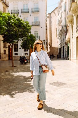 Şık giyinmiş genç bir kadın şehrin güneşli caddelerinde geziniyor. Kıvırcık saçlı güzel bir kadın gün batımını dışarıda izlemekten hoşlanır. Turizm ve yürüme kavramı.