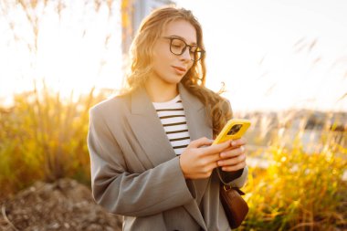 Gözlüklü gülümseyen bir kadın, çatıdaki terasta telefonuyla duruyor ve günbatımının tadını çıkarıyor. Güzel bir kadın dışarıda poz veriyor. Şehir kavramı, moda ve blog yazma.