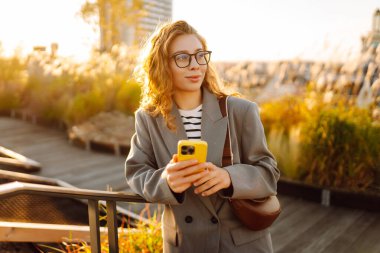 Gözlüklü gülümseyen bir kadın, çatıdaki terasta telefonuyla duruyor ve günbatımının tadını çıkarıyor. Güzel bir kadın dışarıda poz veriyor. Şehir kavramı, moda ve blog yazma.