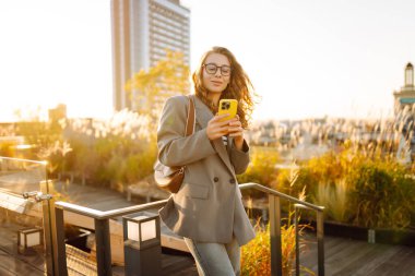 Gözlüklü gülümseyen bir kadın, çatıdaki terasta telefonuyla duruyor ve günbatımının tadını çıkarıyor. Güzel bir kadın dışarıda poz veriyor. Şehir kavramı, moda ve blog yazma.