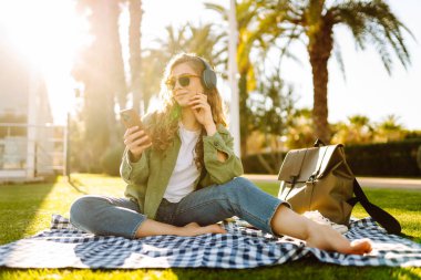 Kablosuz kulaklığı ve telefonu olan genç bir kadın güneşli bir parkta, palmiye ağaçlarıyla çevrili bir battaniyenin üzerinde oturuyor. Güzel kadın eğleniyor, müzik dinliyor ve dışarıda dinleniyor. Yaşam biçimi.