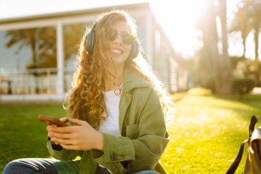 Kablosuz kulaklığı ve telefonu olan genç bir kadın güneşli bir parkta, palmiye ağaçlarıyla çevrili bir battaniyenin üzerinde oturuyor. Güzel kadın eğleniyor, müzik dinliyor ve dışarıda dinleniyor. Yaşam biçimi.