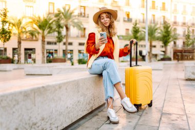 Sarı bavullu ve telefonlu neşeli kadın şehrin mimarisine hayran. Şapkalı ve sırt çantalı bir turist şehrin güneşli caddelerinde geziyor. Turizm kavramları, hafta sonları.