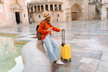 Sarı bavullu ve telefonlu neşeli kadın şehrin mimarisine hayran. Şapkalı ve sırt çantalı bir turist şehrin güneşli caddelerinde geziyor. Turizm kavramları, hafta sonları.