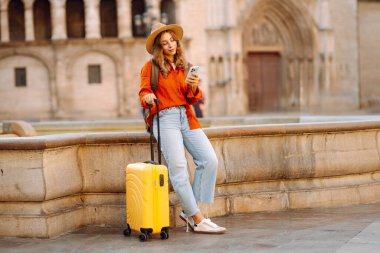 Sarı bavullu ve telefonlu neşeli kadın şehrin mimarisine hayran. Şapkalı ve sırt çantalı bir turist şehrin güneşli caddelerinde geziyor. Turizm kavramları, hafta sonları.