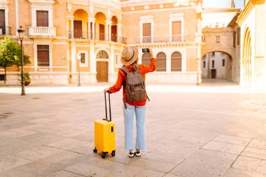 Sarı bavullu ve telefonlu neşeli kadın şehrin mimarisine hayran. Şapkalı ve sırt çantalı bir turist şehrin güneşli caddelerinde geziyor. Turizm kavramları, hafta sonları.