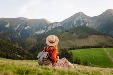Sırt çantalı genç bir kadın gezgin gün batımında yeşil bir tepe boyunca geziniyor. Kadın dağ manzarasından zevk alıyor ve açık havanın özgürlüğünü hissediyor. Doğa, seyahat ve özgürlük kavramları.