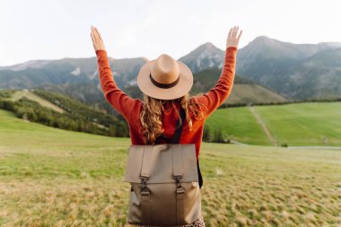 Sırt çantalı genç bir kadın gezgin gün batımında yeşil bir tepe boyunca geziniyor. Kadın dağ manzarasından zevk alıyor ve açık havanın özgürlüğünü hissediyor. Doğa, seyahat ve özgürlük kavramları.