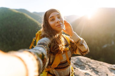Gün batımında arka planda dağ sırasıyla selfie çeken kadın portresi. Sarı sırt çantası blogları olan güzel bir gezgin ve doğada güneşli bir günün tadını çıkarıyor. Macera kavramları, özgürlük, blog yazma.