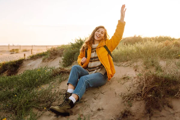 Parlak ceketli genç bir kadın gün batımında kumlu bir sahilde yürüyor. Güzel bir sırt çantalı doğa ve açık hava macerasından zevk alır. Seyahat, doğa ve rahatlama kavramı.