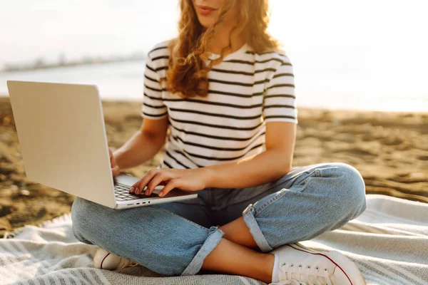 Güneşli bir günde plaj battaniyesinde otururken dizüstü bilgisayarla çalışan serbest çalışan bir kadın. Dizüstü bilgisayarı olan güzel bir kadın gün batımında doğanın ve deniz manzarasının tadını çıkarıyor. Rahatlama kavramı, dinlenme.