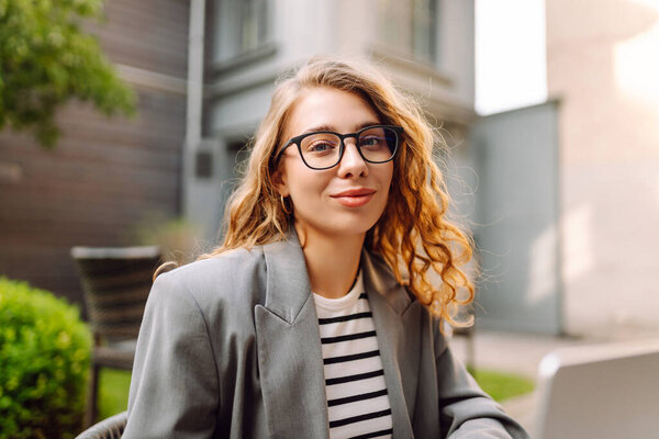 Веселая женщина с ноутбуком сидит за столом на террасе кафе. Женщина-фрилансер работает над своим ноутбуком, пьет кофе и наслаждается солнечным днем. Концепция удаленной работы, выходные.