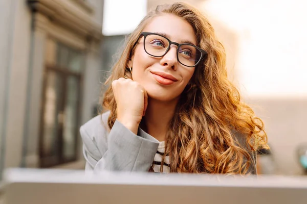 Dizüstü bilgisayarı olan neşeli bir kadın kafe terasında bir masada oturuyor. Dişi bir serbest çalışan dizüstü bilgisayarında çalışır, kahve içer ve güneşli günün tadını çıkarır. Uzaktan çalışma konsepti, hafta sonu.