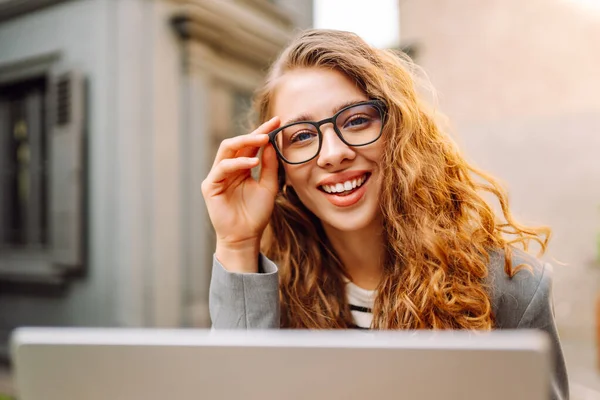 Dizüstü bilgisayarı olan neşeli bir kadın kafe terasında bir masada oturuyor. Dişi bir serbest çalışan dizüstü bilgisayarında çalışır, kahve içer ve güneşli günün tadını çıkarır. Uzaktan çalışma konsepti, hafta sonu.