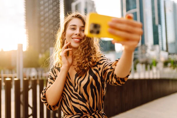 Mutlu bir kadın telefonuyla modern bir şehrin arka planına selfie çeker. Telefon blogları olan güzel bir turist ve gün batımının tadını çıkarıyor. Blogculuk, boş zaman ve kentsellik kavramları.