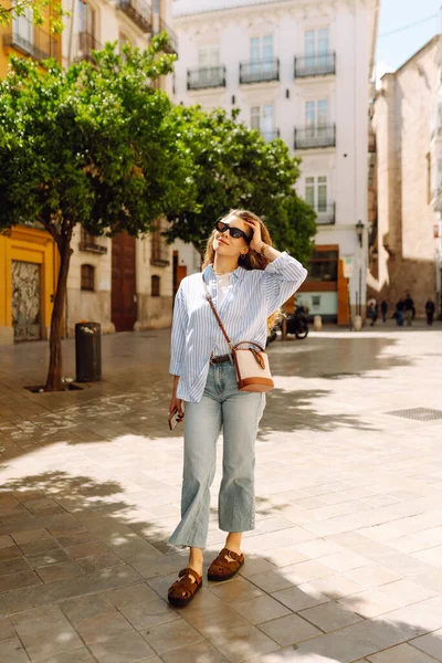 Şık giyinmiş genç bir kadın şehrin güneşli caddelerinde geziniyor. Kıvırcık saçlı güzel bir kadın gün batımını dışarıda izlemekten hoşlanır. Turizm ve yürüme kavramı.