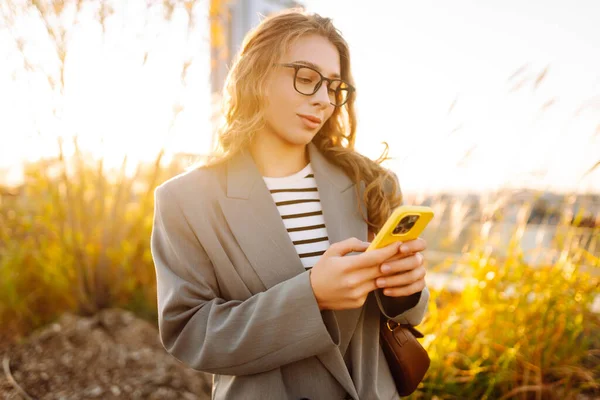 Gözlüklü gülümseyen bir kadın, çatıdaki terasta telefonuyla duruyor ve günbatımının tadını çıkarıyor. Güzel bir kadın dışarıda poz veriyor. Şehir kavramı, moda ve blog yazma.