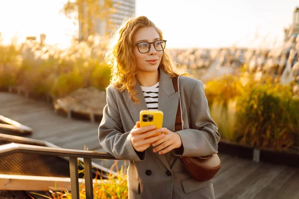 Gözlüklü gülümseyen bir kadın, çatıdaki terasta telefonuyla duruyor ve günbatımının tadını çıkarıyor. Güzel bir kadın dışarıda poz veriyor. Şehir kavramı, moda ve blog yazma.
