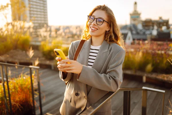 Gözlüklü gülümseyen bir kadın, çatıdaki terasta telefonuyla duruyor ve günbatımının tadını çıkarıyor. Güzel bir kadın dışarıda poz veriyor. Şehir kavramı, moda ve blog yazma.