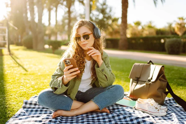 Kablosuz kulaklığı ve telefonu olan genç bir kadın güneşli bir parkta, palmiye ağaçlarıyla çevrili bir battaniyenin üzerinde oturuyor. Güzel kadın eğleniyor, müzik dinliyor ve dışarıda dinleniyor. Yaşam biçimi.